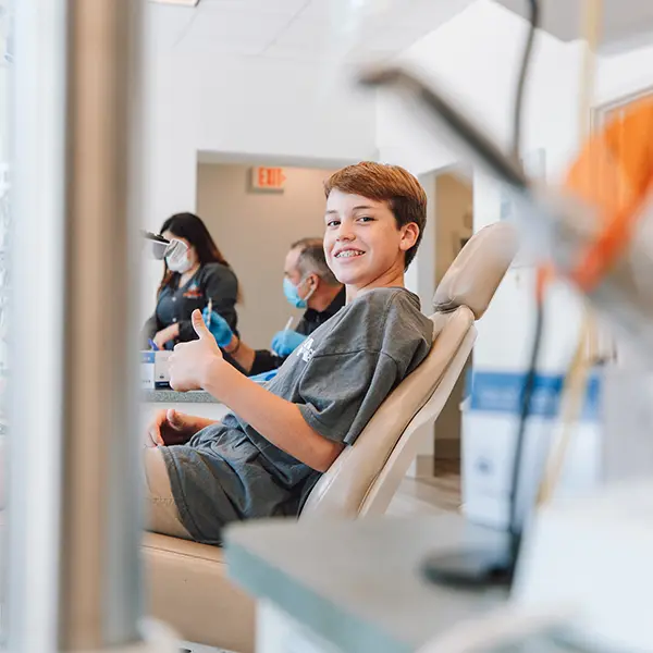 Caucasian teenage boy sitting an orthodontist chair, smiling with a thumbs-up.